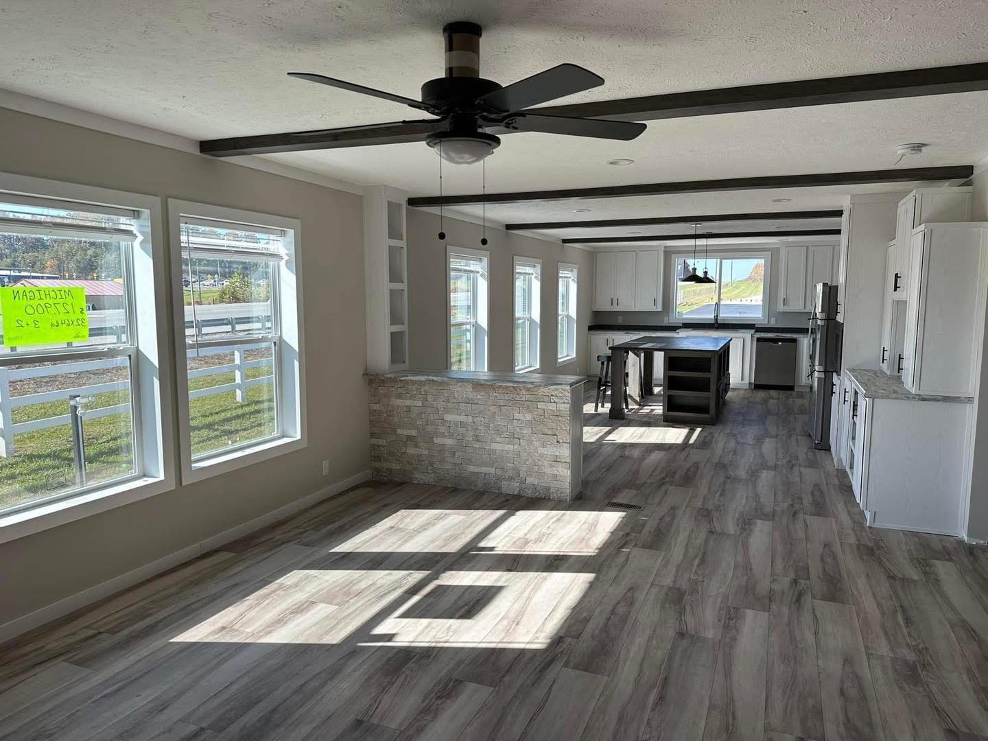 Open, modern kitchen with large windows, stone island, wood floors, and a ceiling fan. Sunlight streams in, creating a bright and airy ambiance.