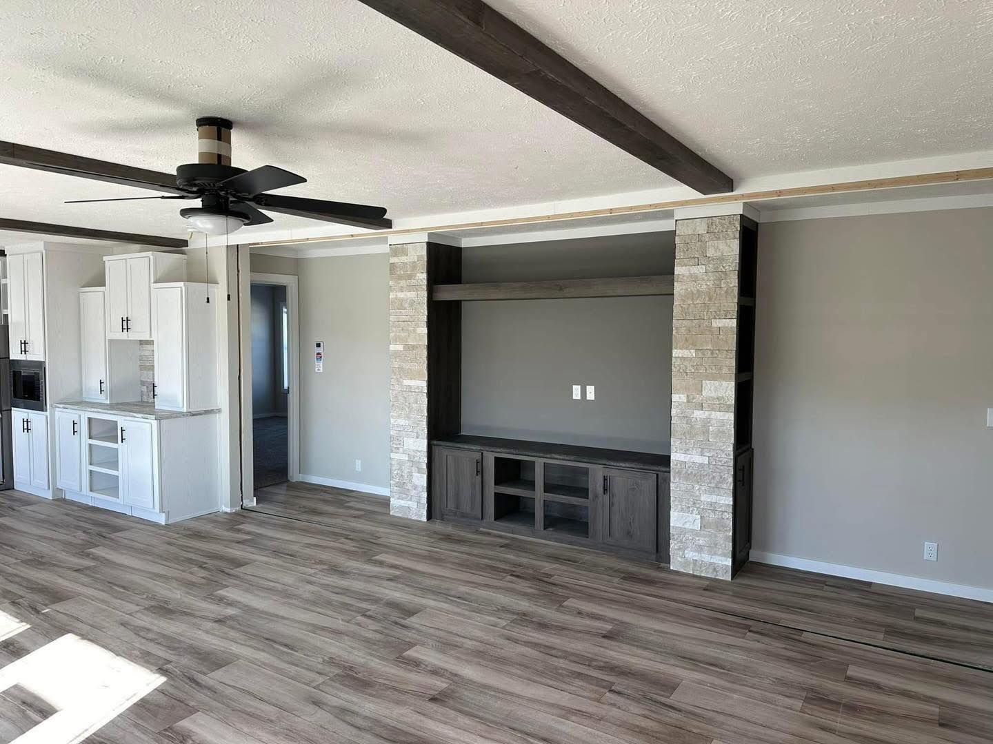 Spacious living area with light wood flooring, a dark wood entertainment center flanked by stone columns, and a ceiling fan. Modern open layout, bright and neutral.