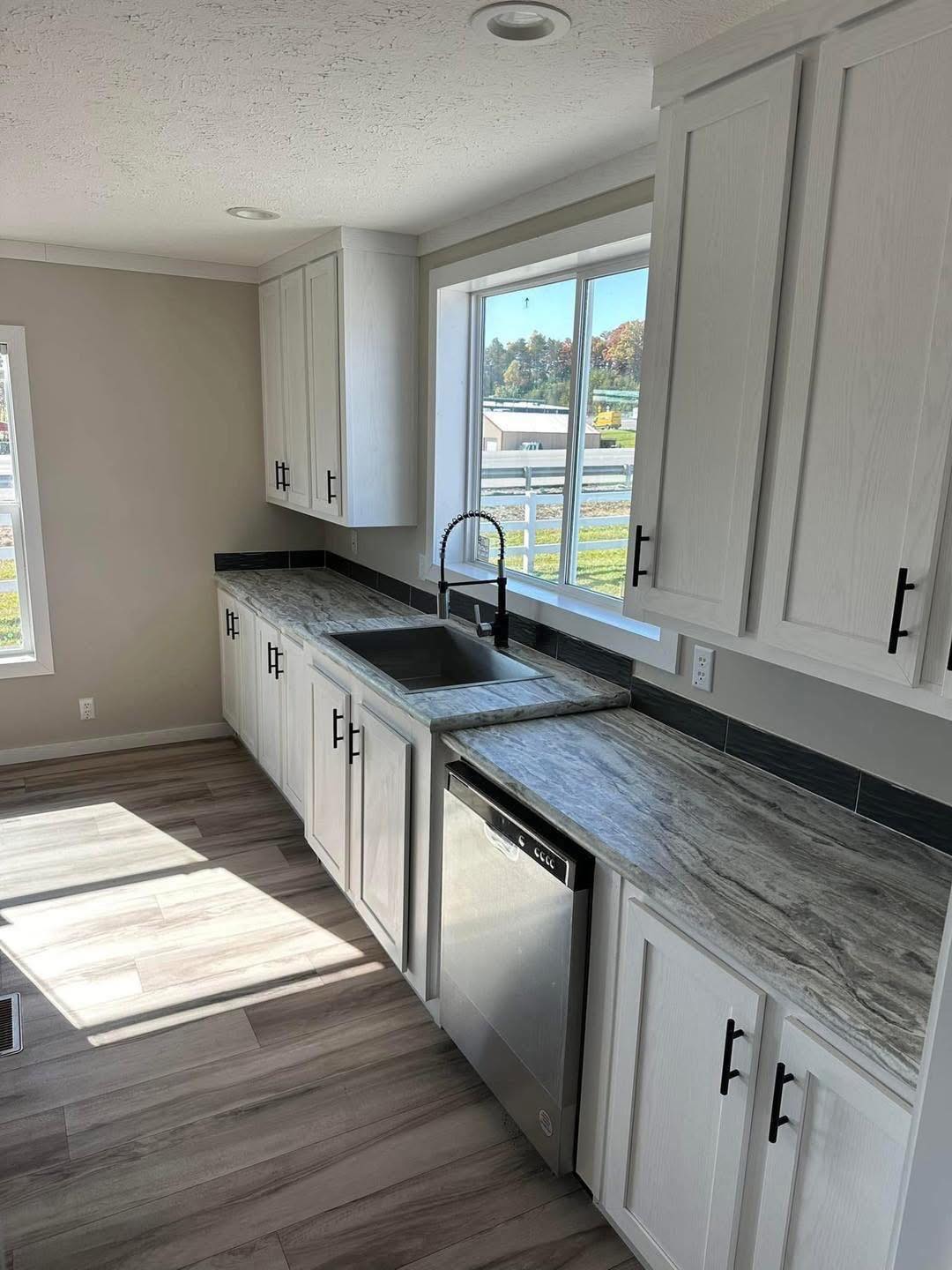 Modern kitchen with white cabinets, marble countertops, and a stainless steel dishwasher. Light floods through a large window, creating an airy feel.