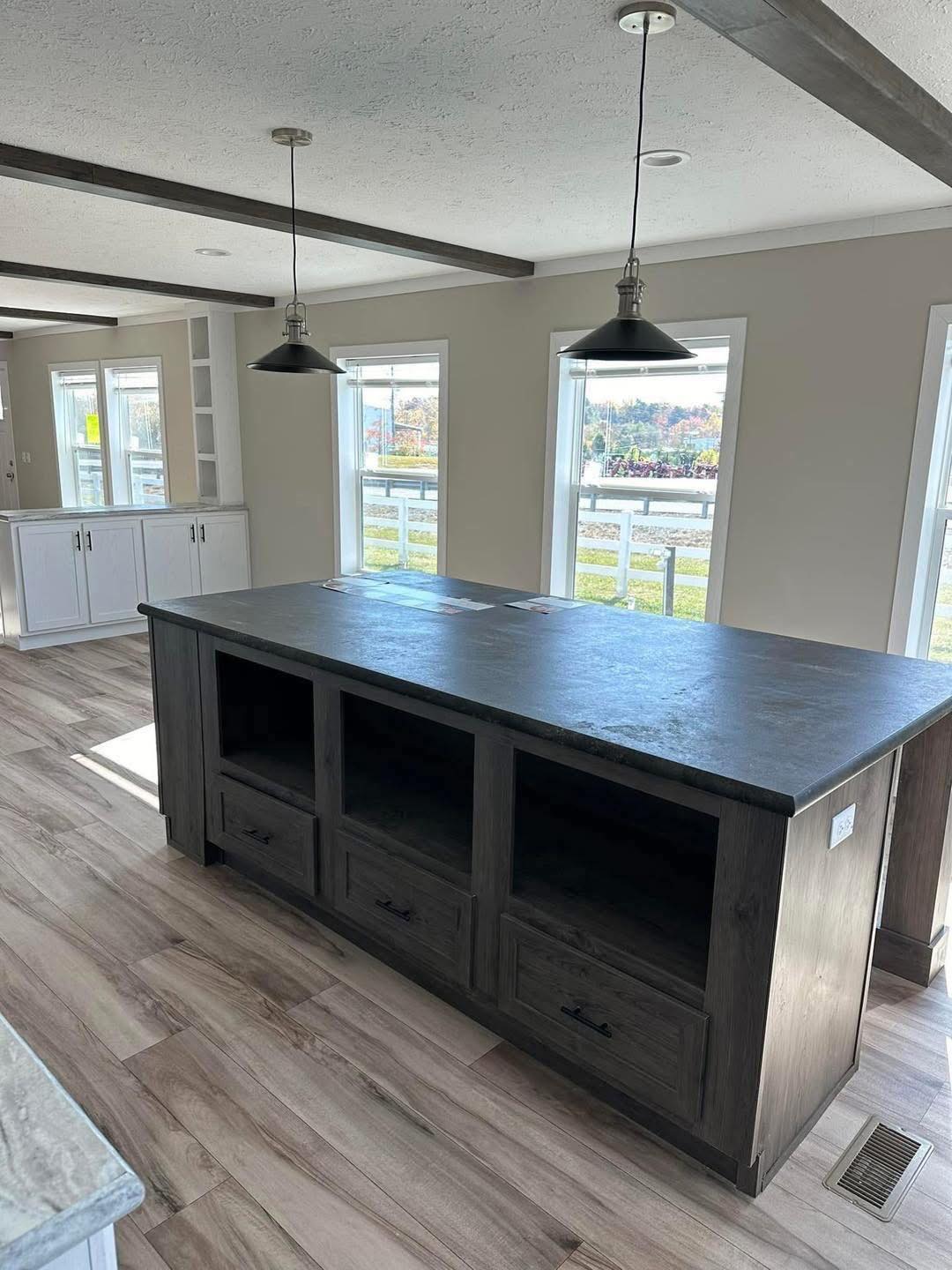 Spacious kitchen with a dark wooden island and three drawers, illuminated by pendant lights. Large windows offer ample natural light and a view outside.