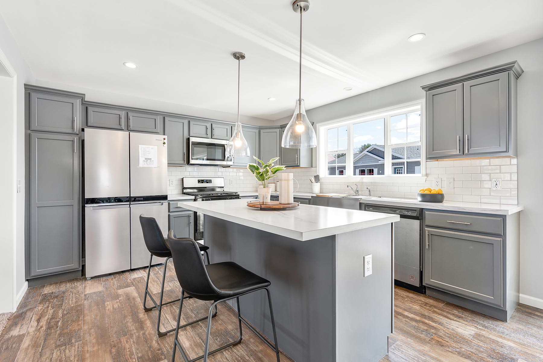 Modern kitchen with gray cabinets, stainless steel appliances, and a large island with pendant lights. Bright and airy with wooden floors and a window view.