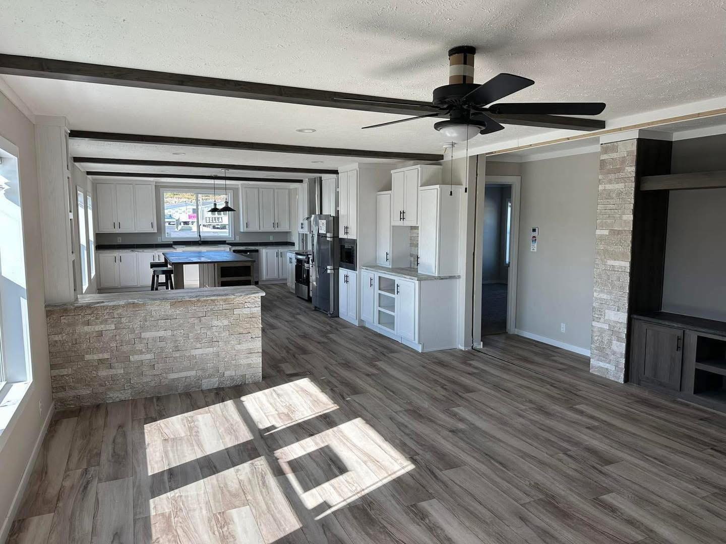 Spacious modern kitchen and living area with light wood floors, white cabinetry, and a stone island. Large windows and ceiling fan illuminate the space.