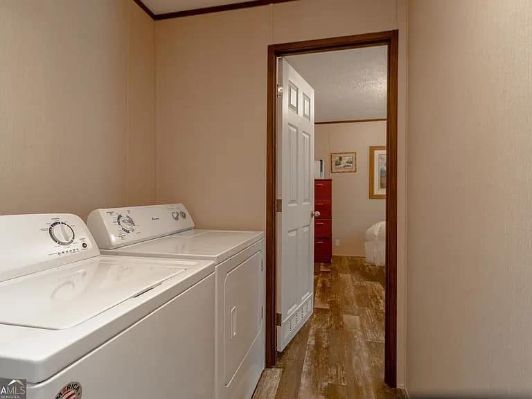 Laundry room with beige walls, featuring a white washer and dryer on the left. Open door leads to a bedroom with wooden flooring and framed artwork.