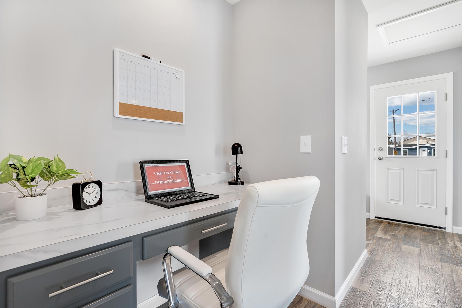 Modern home office with a white desk and gray drawers. A laptop, lamp, clock, and plant sit on the desk. A calendar hangs above. Bright, minimal, and organized space.