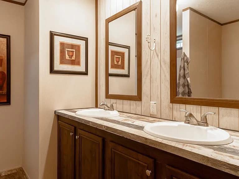 Rustic bathroom with dual sinks on a wooden counter, flanked by mirrors. Earth-toned artwork hangs on light paneled walls; soft, warm ambiance.