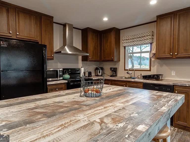 Rustic kitchen with dark wood cabinets, a black fridge, and a stainless steel range hood. A distressed wooden island holds a wire basket. Cozy and inviting.