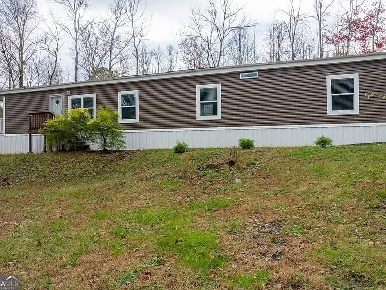 A long, brown mobile home with white trim sits on a grassy, slightly sloped yard. Leafless trees stand behind under a cloudy sky, creating a serene, rustic setting.