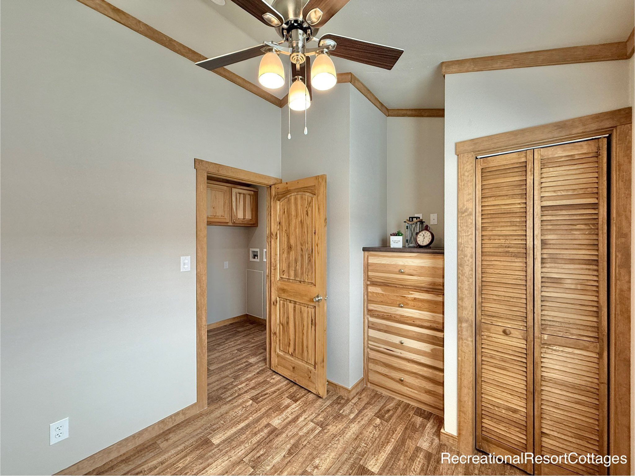 Cozy room with wooden floors, light gray walls, and wooden trim. Open door leads to a utility area. Ceiling fan and rustic dresser add warmth.