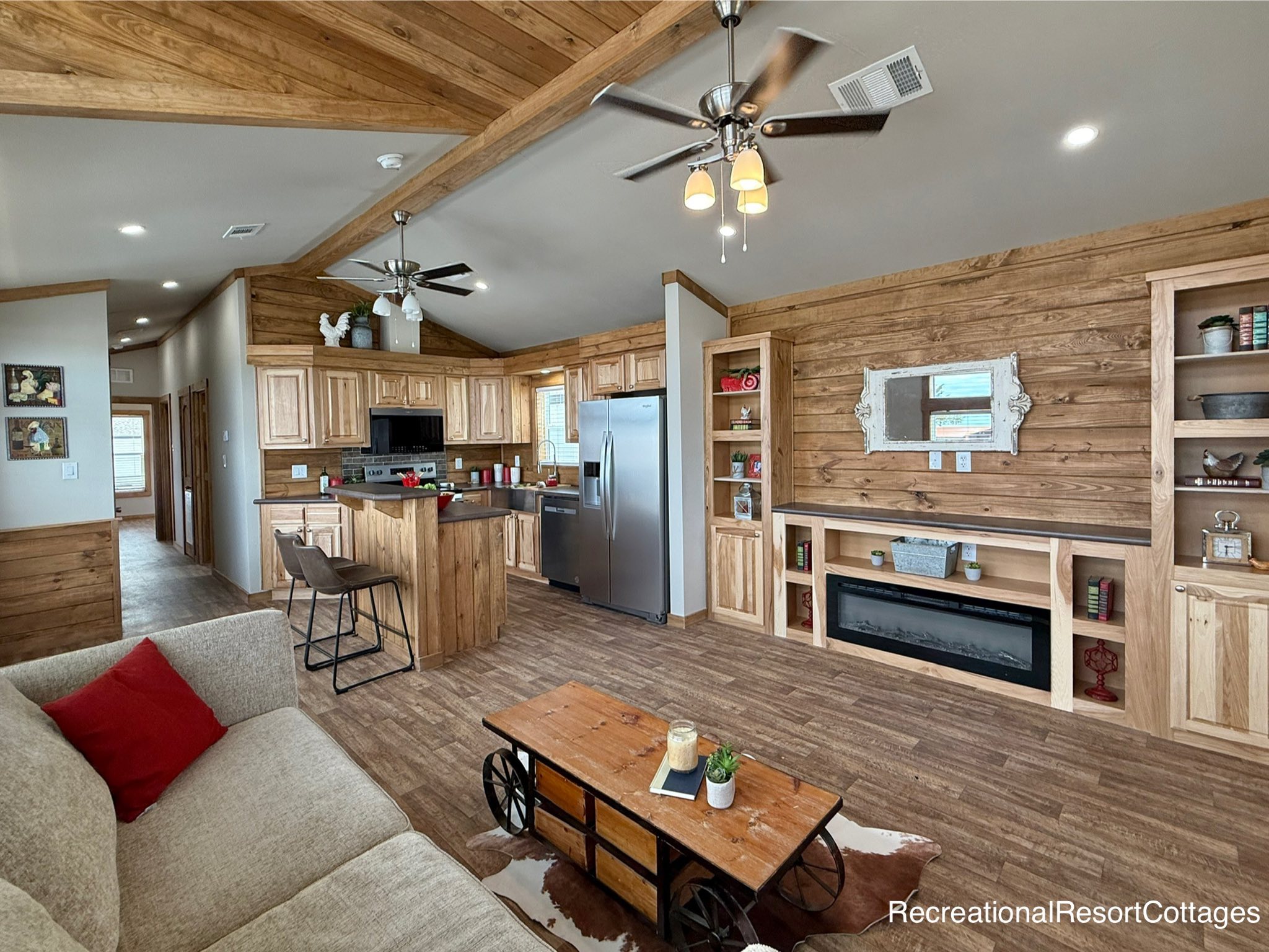 Rustic living room and kitchen with wooden accents, ceiling fans, and cozy seating. Modern appliances are featured in the open-concept space. Warm and inviting.