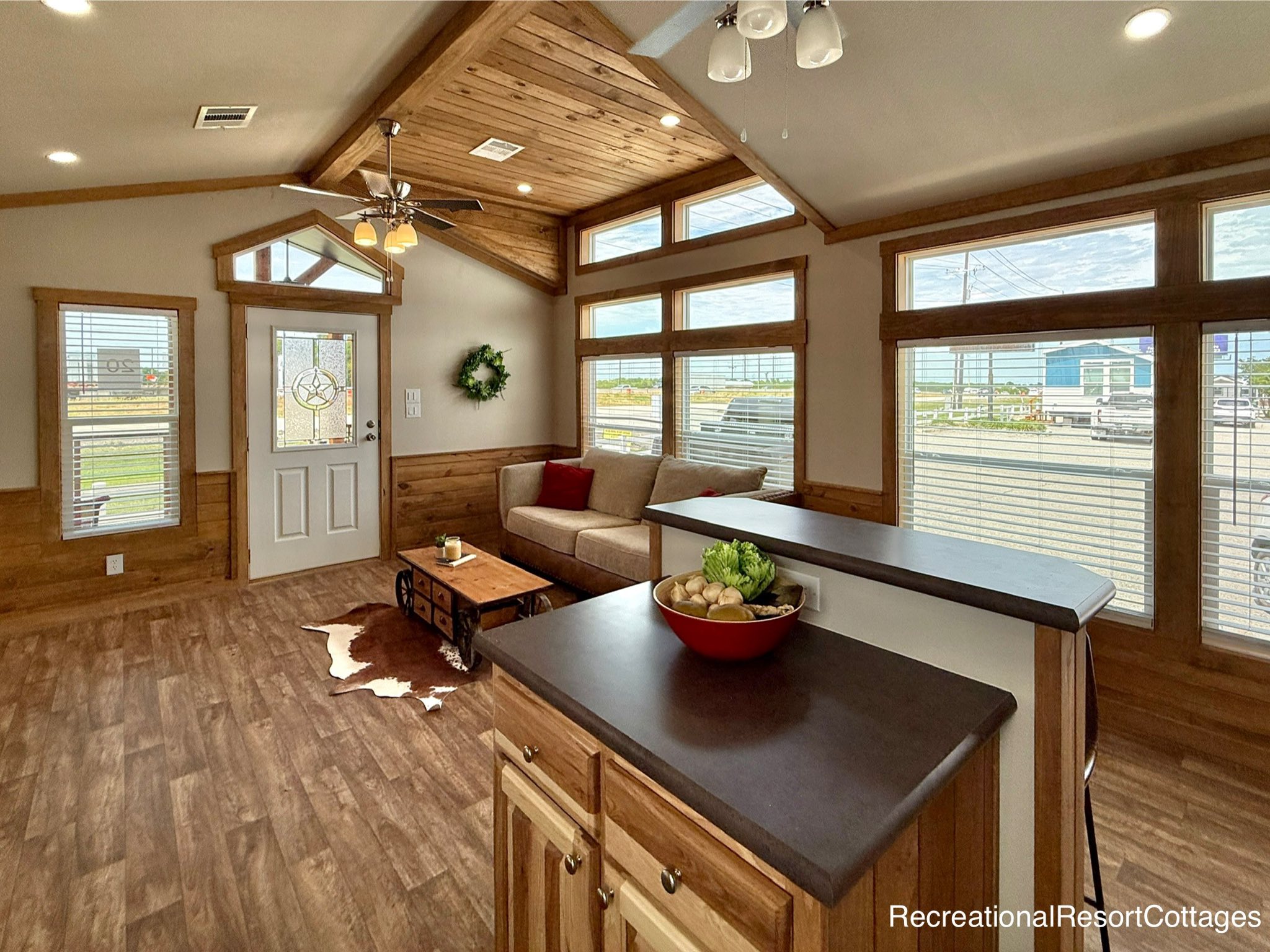 Rustic living room with wood accents, high ceilings, and large windows. Features a beige sofa, cowhide rug, and kitchen island with fruit bowl.