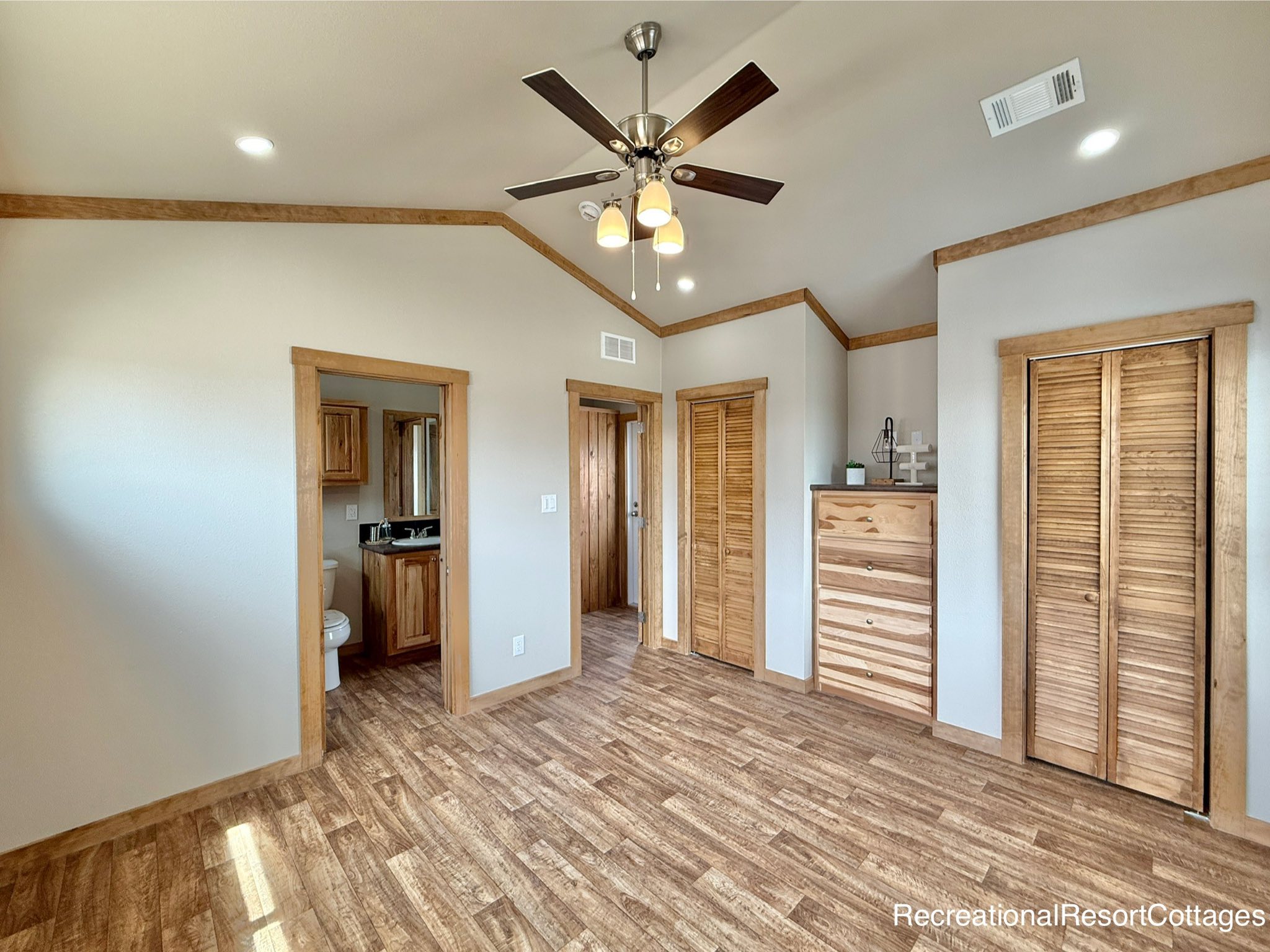 A cozy room with wood accents features a ceiling fan, light wooden floors, a dresser, and louvered closet doors. The area feels warm and inviting.