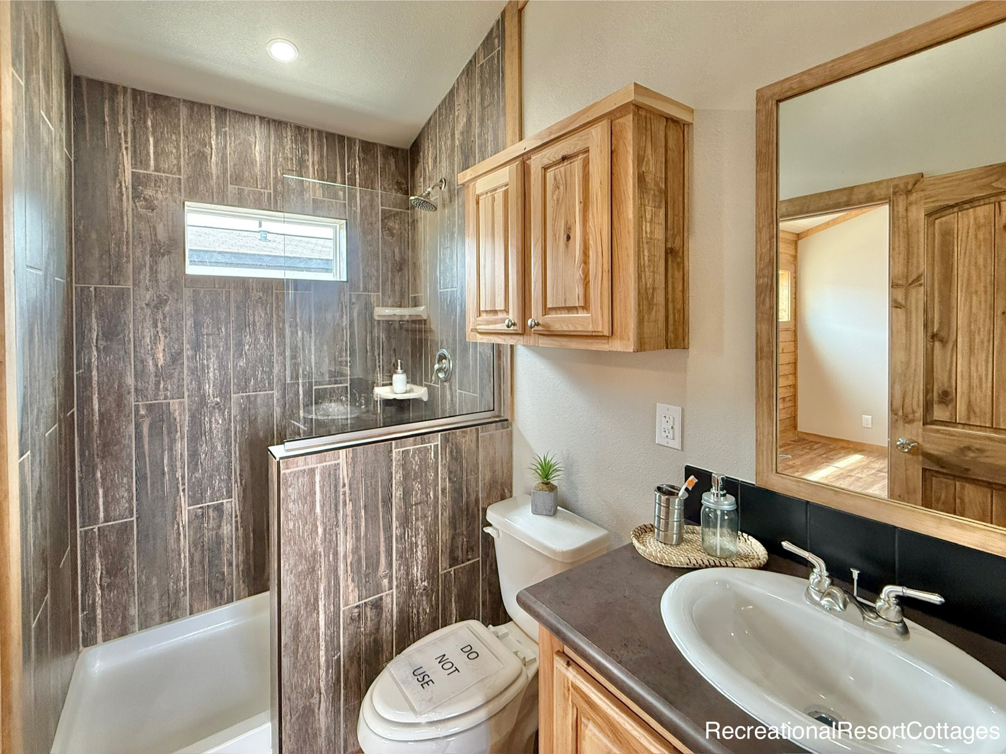 Compact bathroom with rustic wooden cabinets and dark brown tiled shower. White sink with silver faucet and a plant next to a sign on the toilet lid.