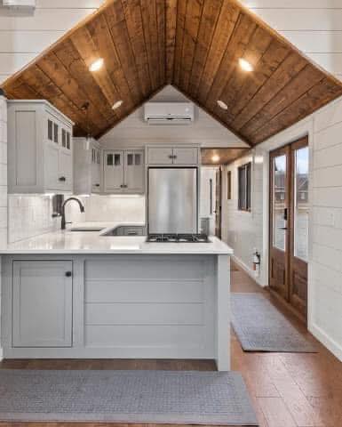 A cozy kitchen with white cabinets, a wood-paneled cathedral ceiling, and stainless steel appliances. Bright lighting and large windows create a warm, inviting atmosphere.