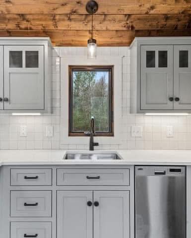 A cozy kitchen with light gray cabinets, a stainless steel dishwasher, and a central window showing a forest view. Features a wooden ceiling and white tiles.