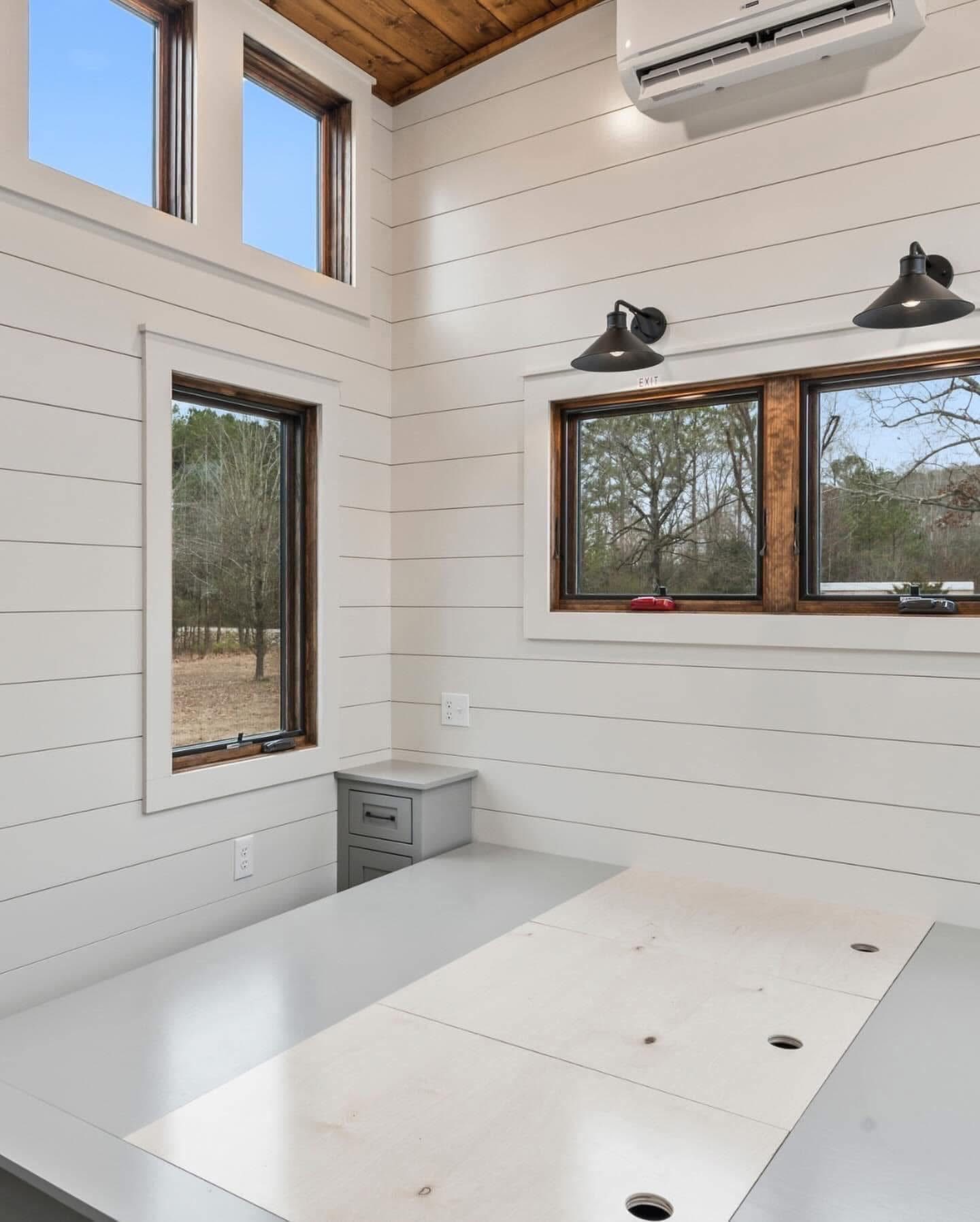 Modern, minimalist room with white shiplap walls, wood ceiling, and large windows. A gray air conditioning unit and desk convey a clean, serene feel.
