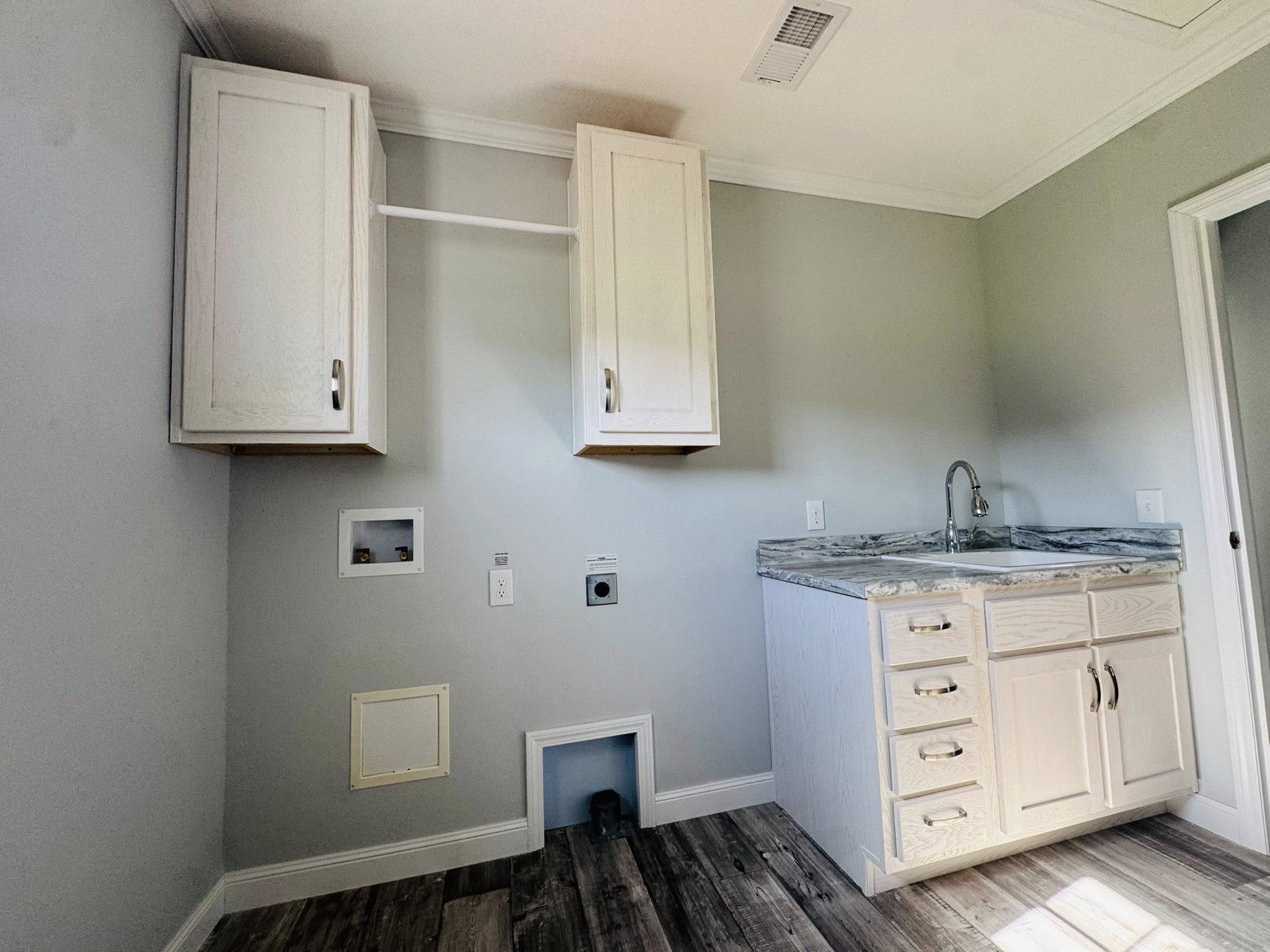 A laundry room with light gray walls and wood flooring. Two white cabinets are mounted on the wall above hookups for laundry appliances. A sink with a marble countertop and white drawers is positioned to the right, under a bright light. The mood is clean and organized.
