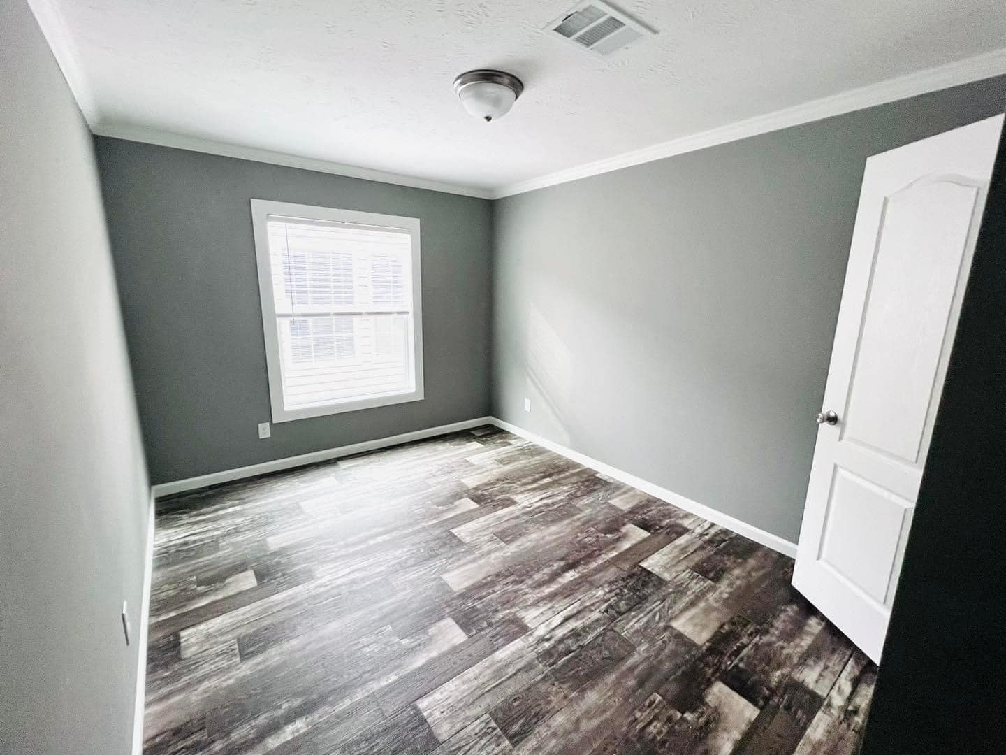 Empty room with gray walls, wooden flooring, and a white ceiling. A closed white door is on the right and a window with blinds lets in natural light.