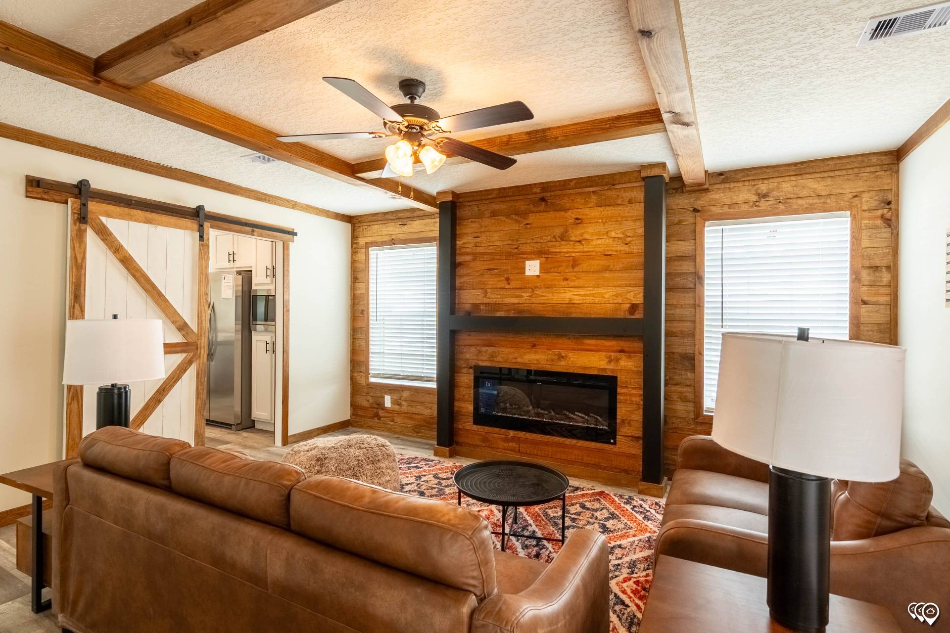 Cozy living room with leather sofas, a colorful rug, and a wooden accent wall featuring a fireplace. A ceiling fan and barn door add rustic charm.