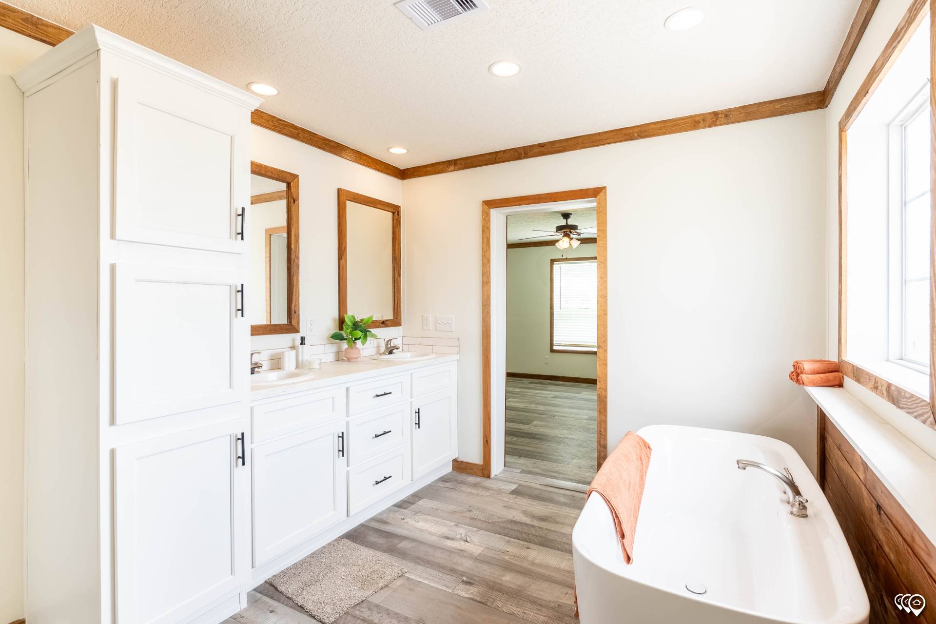 Bright bathroom with white cabinets, dual sinks, and wooden accents. A freestanding tub with an orange towel sits near a large window, creating a fresh and inviting atmosphere.