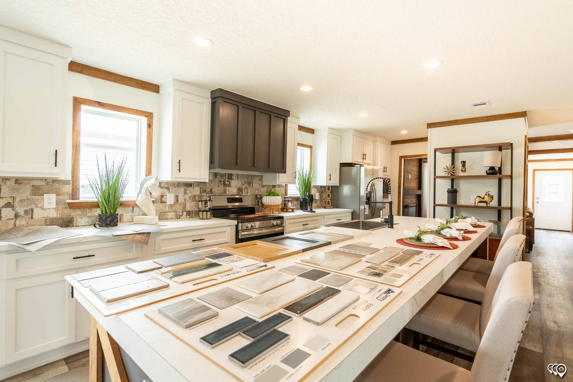 Modern kitchen with white cabinets, centered island displaying tile samples. Light tones, stone backsplash, green plants, and soft natural lighting create an inviting atmosphere.