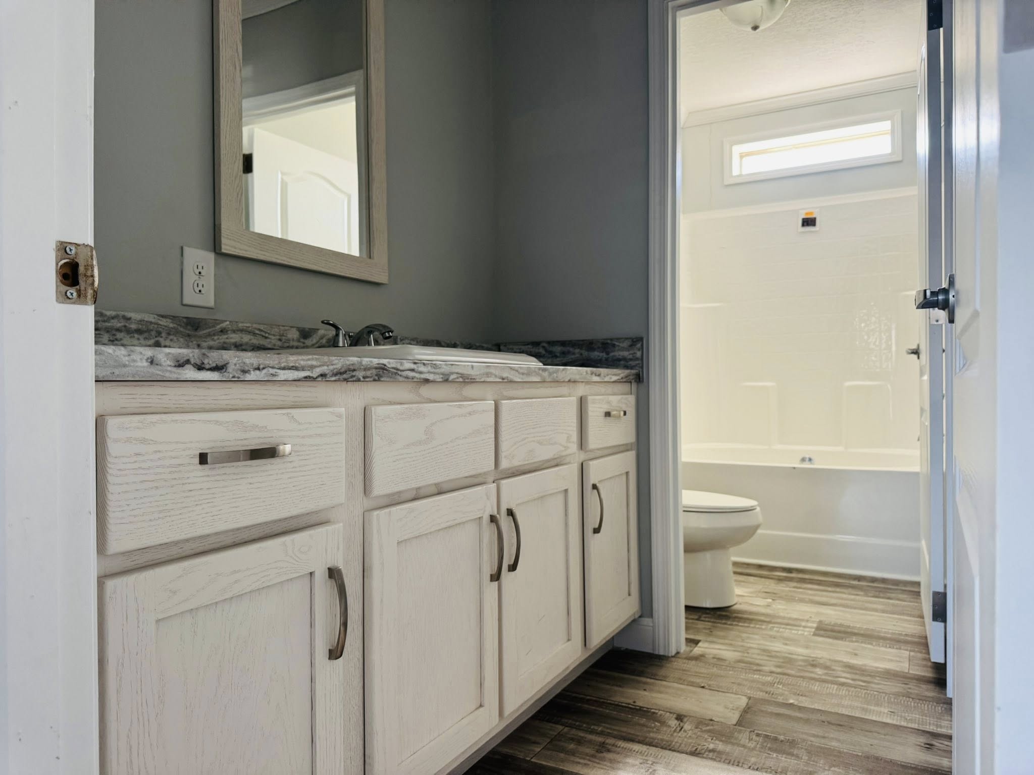 A modern bathroom features light wood cabinets with silver handles, a marble countertop, and a mirror. The adjoining area shows a white bathtub and toilet, with neutral tones and a calm ambiance.