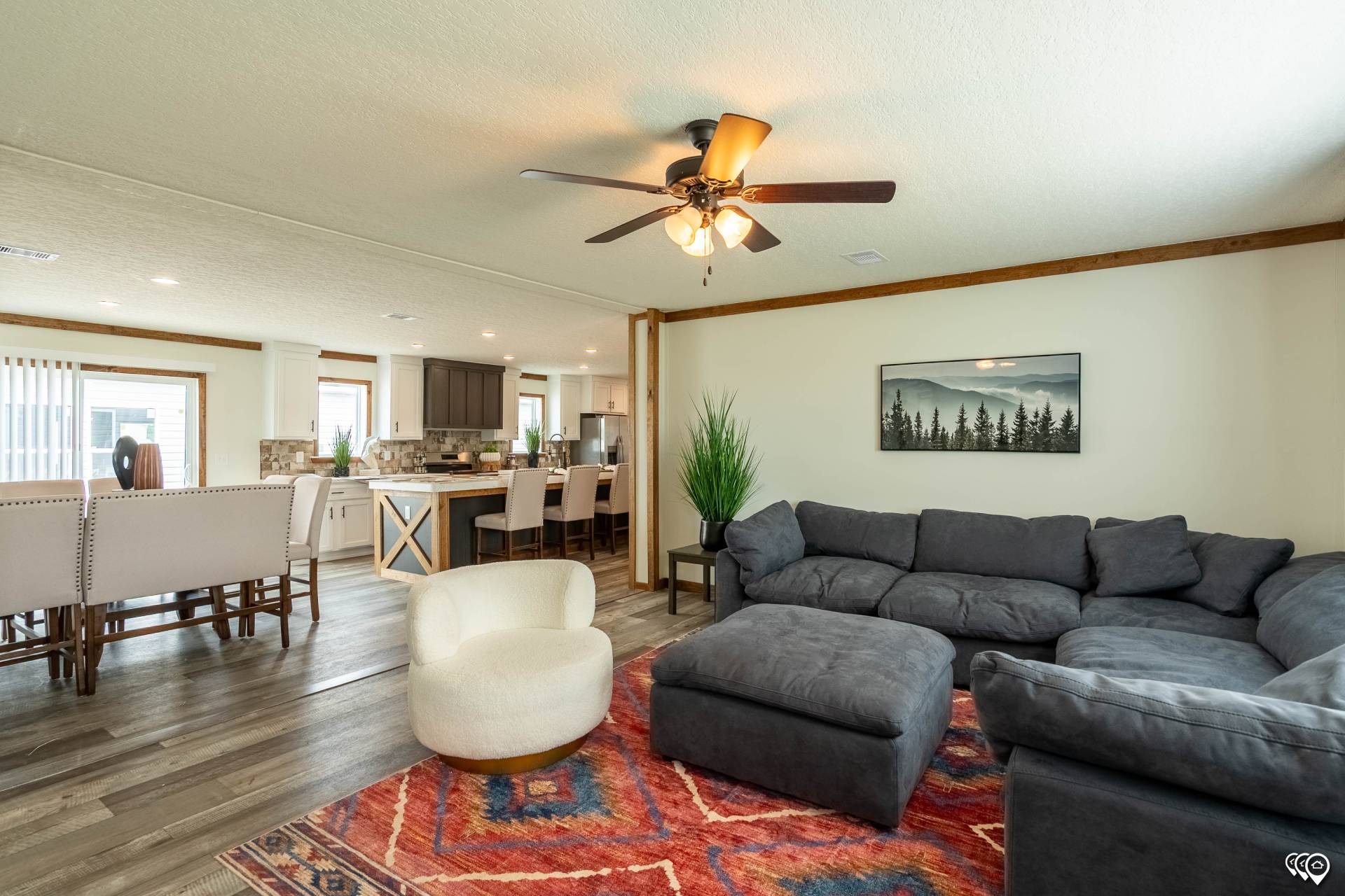 Spacious living room with a dark gray sectional sofa, white swivel chair, and vibrant red patterned rug. A ceiling fan adds warmth. Open kitchen and dining area in background.