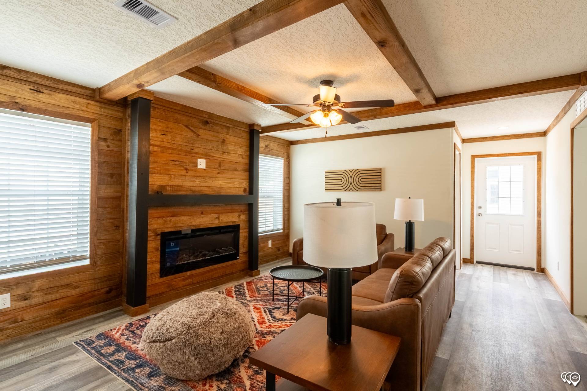 A cozy living room with wooden walls and beams, featuring a brown leather sofa, a modern fireplace, patterned rug, and large windows for natural light.