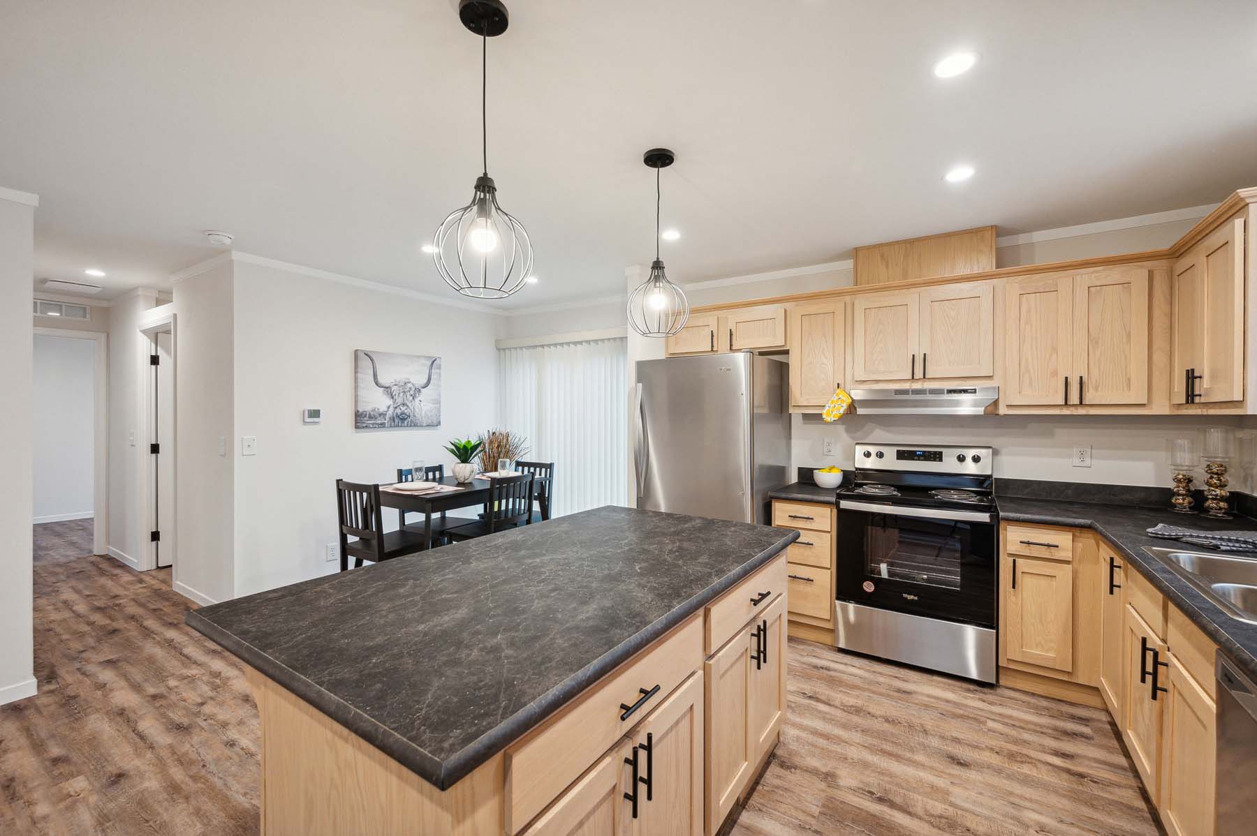 Modern kitchen with wooden cabinets, black countertops, and stainless steel appliances. Two pendant lights hang above a dark island. Dining table in the background with wall art. Warm, inviting tone.