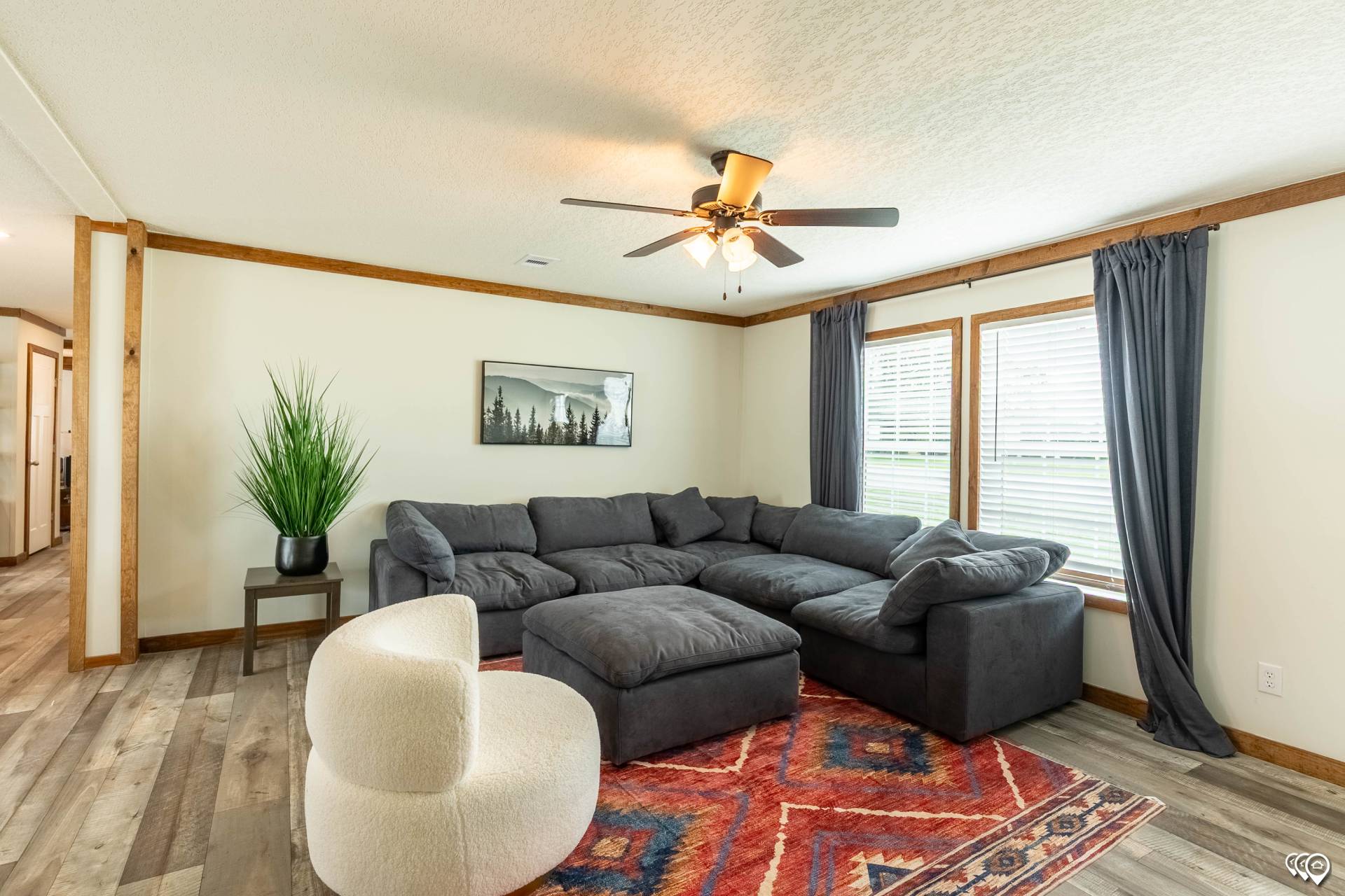 A cozy living room with a large grey sectional sofa, a white rounded chair, and a vibrant red geometric rug. A ceiling fan, potted plant, and woodland artwork enhance the warm, inviting atmosphere.