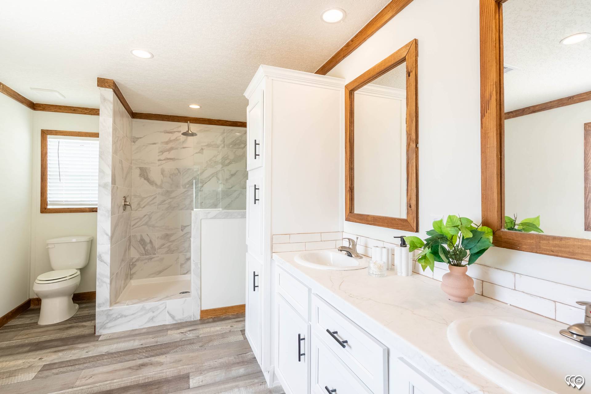 Bright bathroom with white cabinetry and wood accents, featuring a marble-tiled shower, dual sinks with potted plant, and a wooden floor.