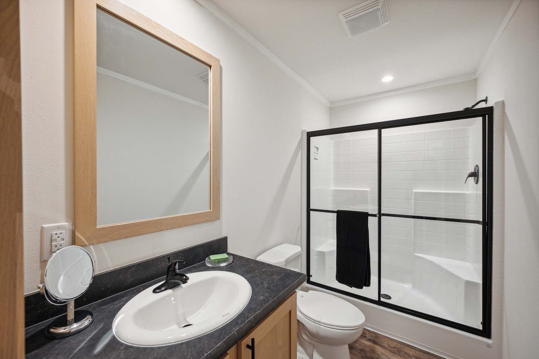 A modern bathroom with a white sink, black countertop, and wooden-framed mirror. A shower with glass doors is in the background, creating a clean, minimalist look.