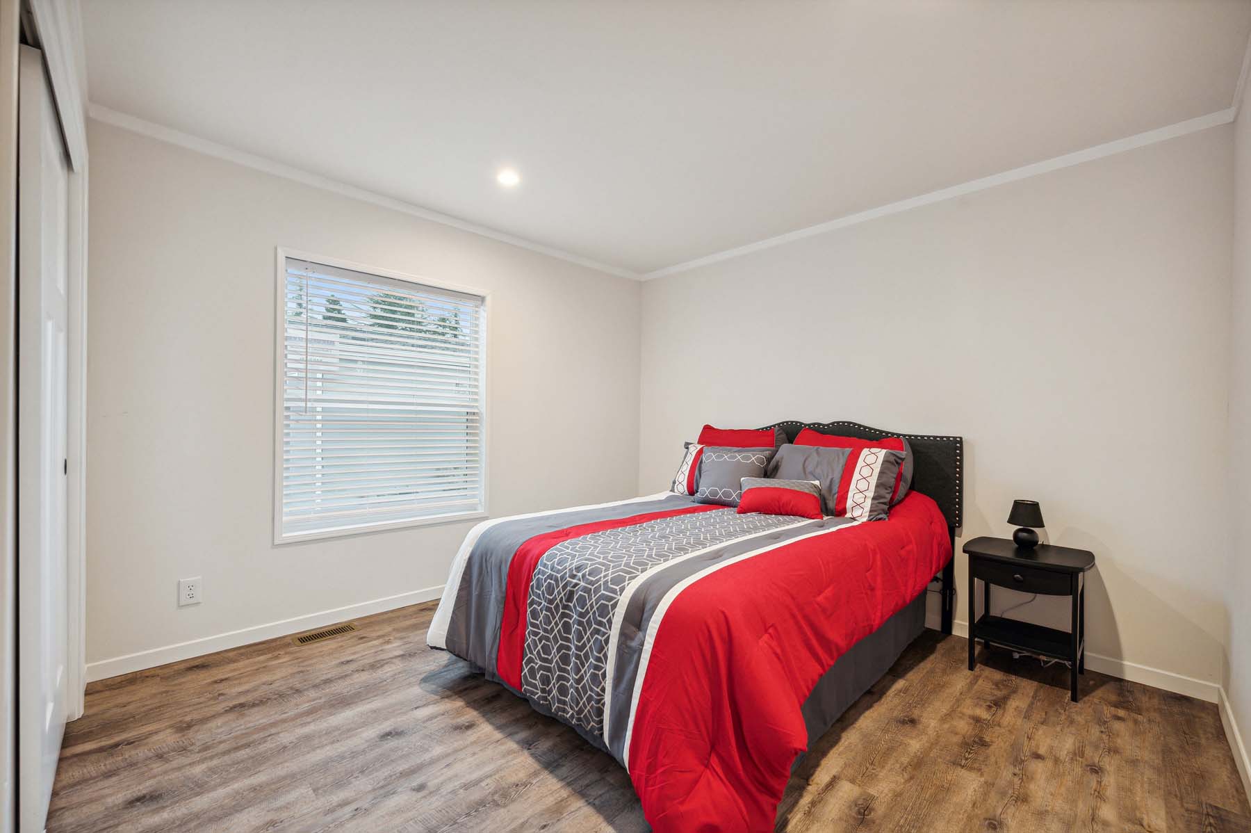 A minimalist bedroom with a neatly made bed covered in red, gray, and white bedding. A window with blinds lets in natural light. There's a small black nightstand with a lamp beside the bed, and the room features wooden flooring, creating a clean and cozy atmosphere.