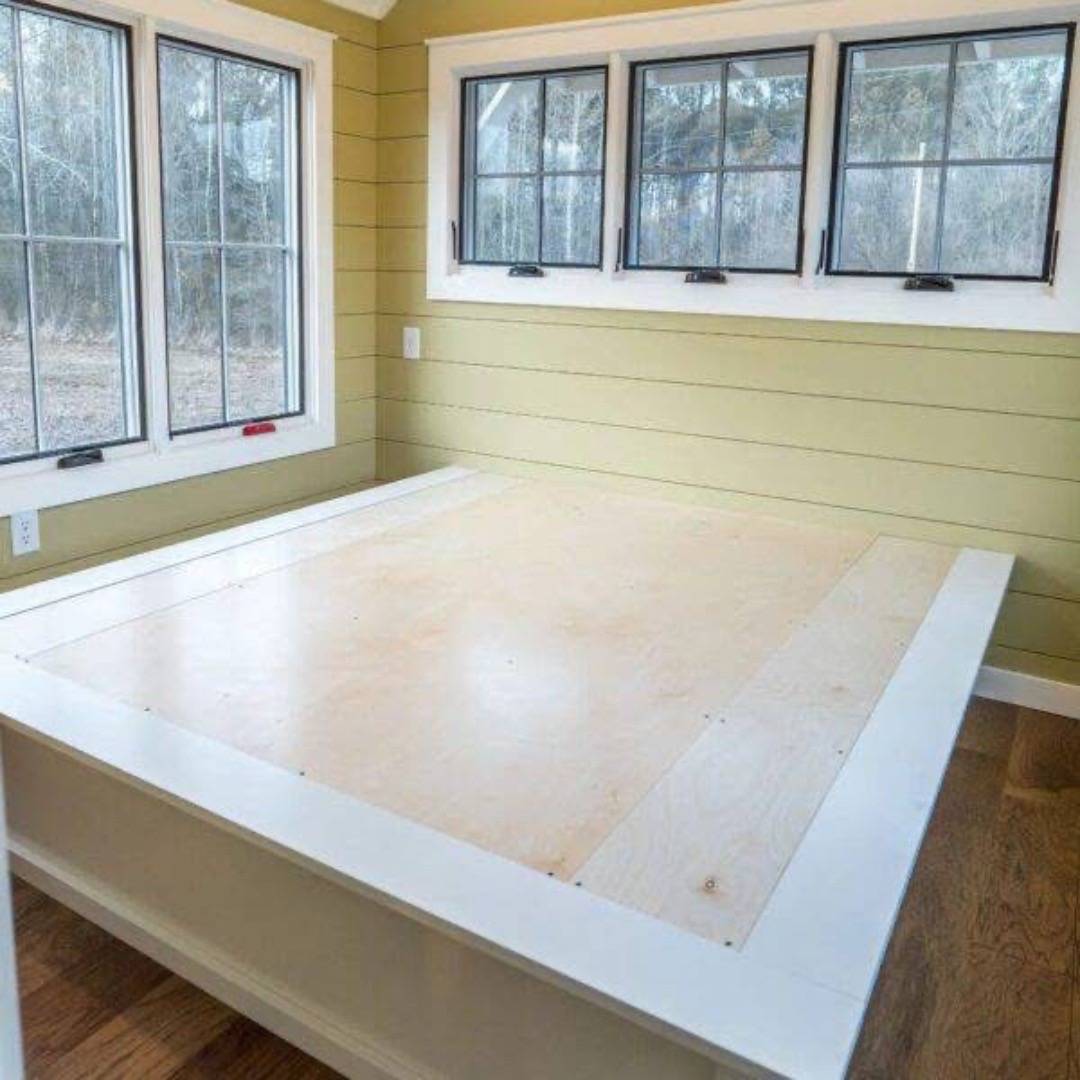 A cozy room features a wooden platform bed with a light wood top and white frame. It is surrounded by green walls and three large windows, creating a bright and inviting atmosphere.