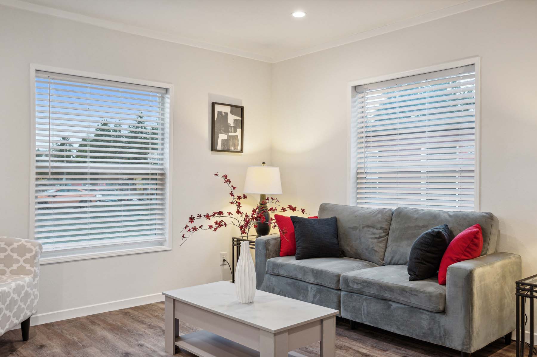 A cozy living room with a gray sofa, adorned with red and black cushions. Two windows with blinds brighten the space, which includes a lamp, decorative branches, and a beige chair.