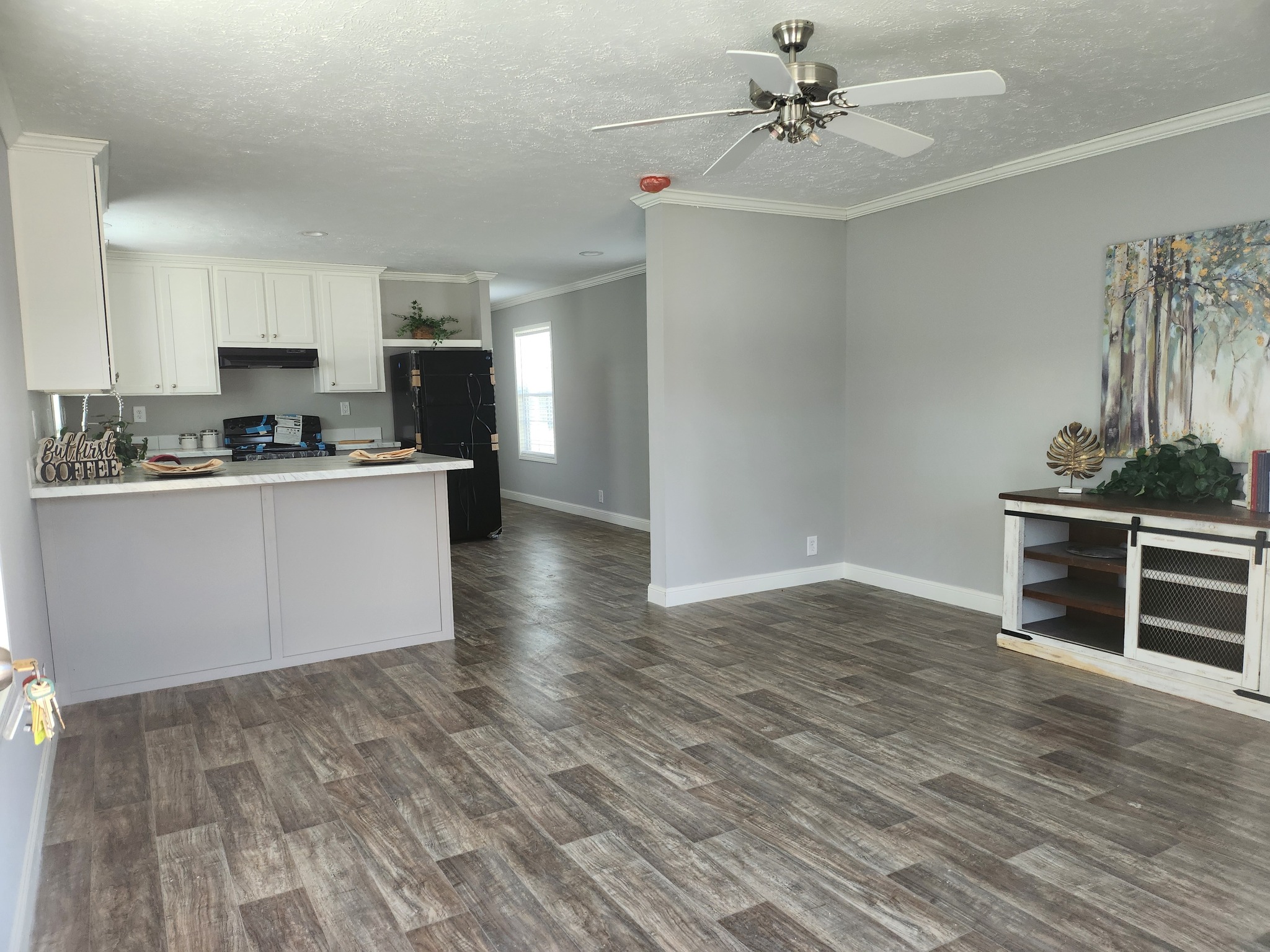 Modern open-plan living area with gray walls, wood floors, and a ceiling fan. Kitchen has white cabinets, a black fridge, and bar counter. Cozy and spacious.