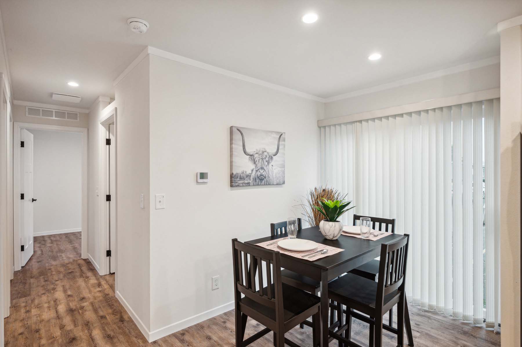 Cozy dining area with a dark wooden table set for four, adorned with a white floral centerpiece. Neutral walls feature a bull artwork. Bright natural light filters through vertical blinds on the right. The wood-textured floor adds warmth, connecting to a hallway on the left, leading to white doors.