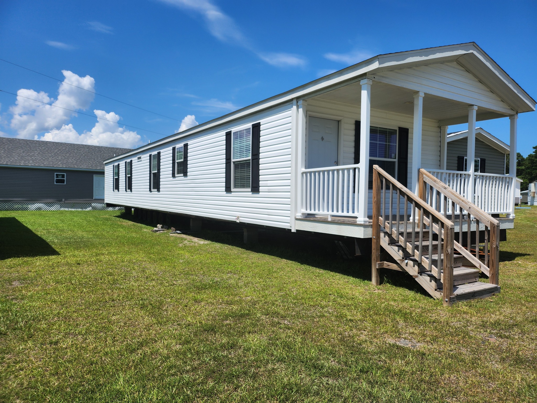 A white mobile home with a small porch and wooden steps sits on a grass lawn under a clear blue sky, conveying a simple, serene suburban feel.