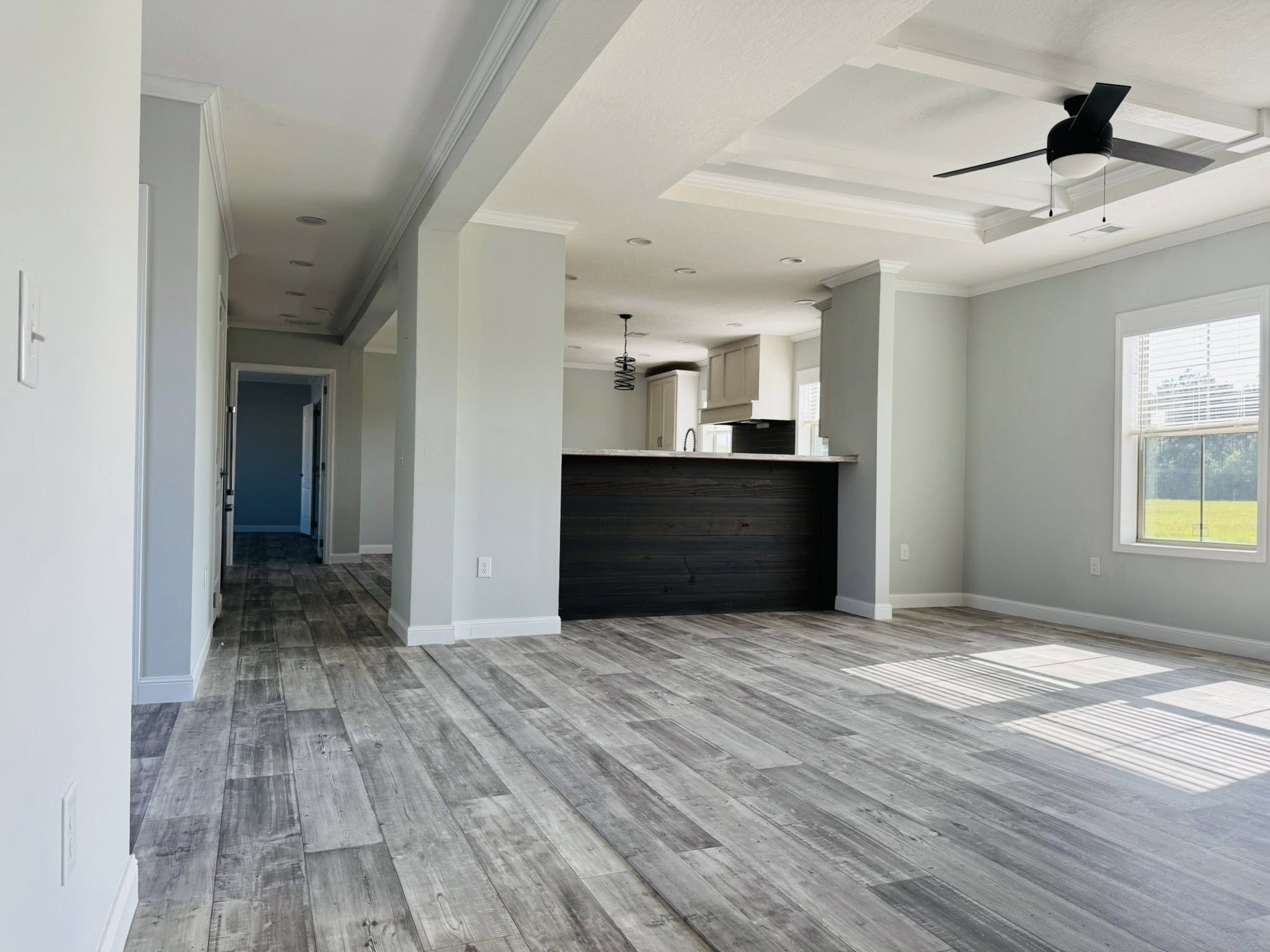 Modern, empty living room with light gray walls, wood floors, and a ceiling fan. Sunlight streams through large windows, creating a bright atmosphere.