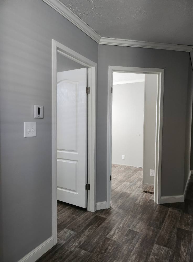 Hallway with two open white doors leading into bright, empty rooms. Gray walls and wood-textured flooring create a clean, modern atmosphere.