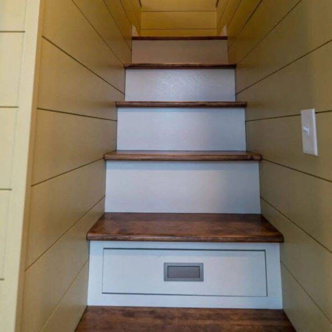 Wooden stairs with light brown paneling on the walls lead upwards. A drawer is built into the lowest step, suggesting a cozy, space-saving design.