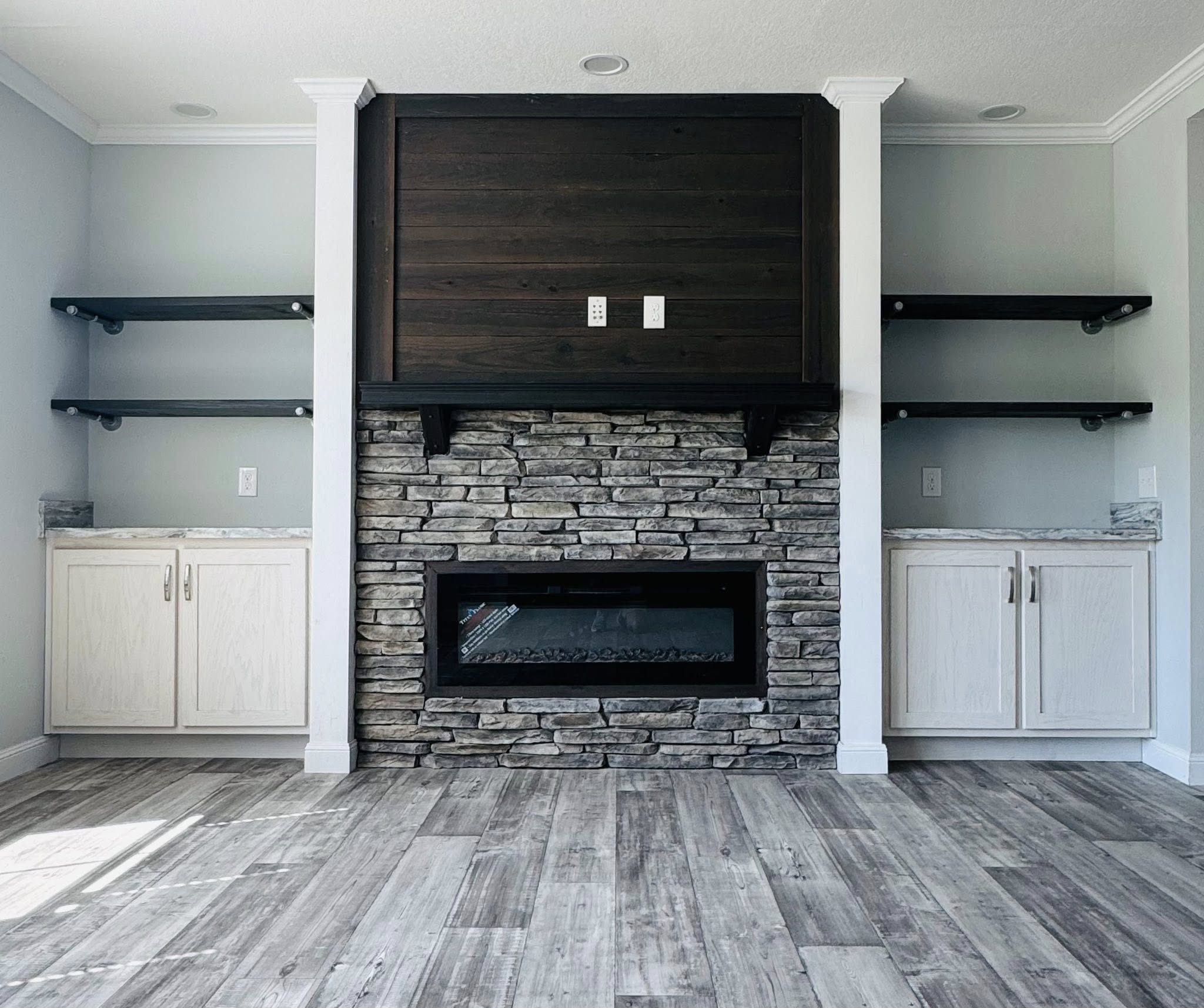 Modern living room with a stone fireplace, dark wooden mantel, and electric insert. White cabinets with gray countertops and dark shelves flank the fireplace.