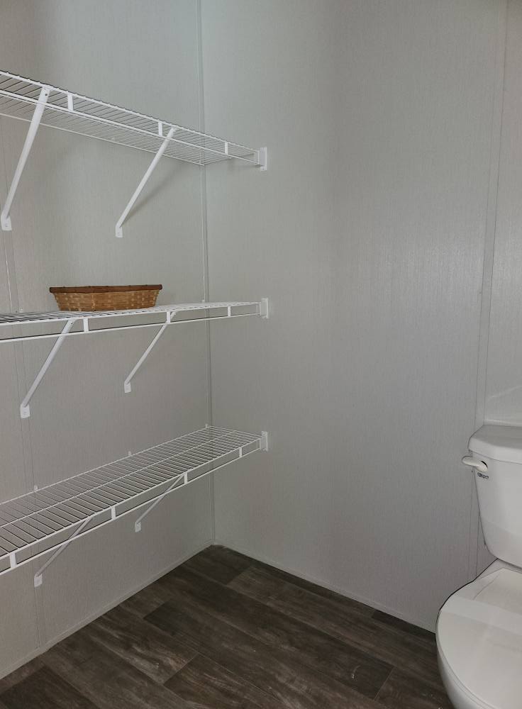 Small bathroom with wire shelves on a pale wall, one holding a rectangular basket. A white toilet is on the right, and the floor is dark wood.