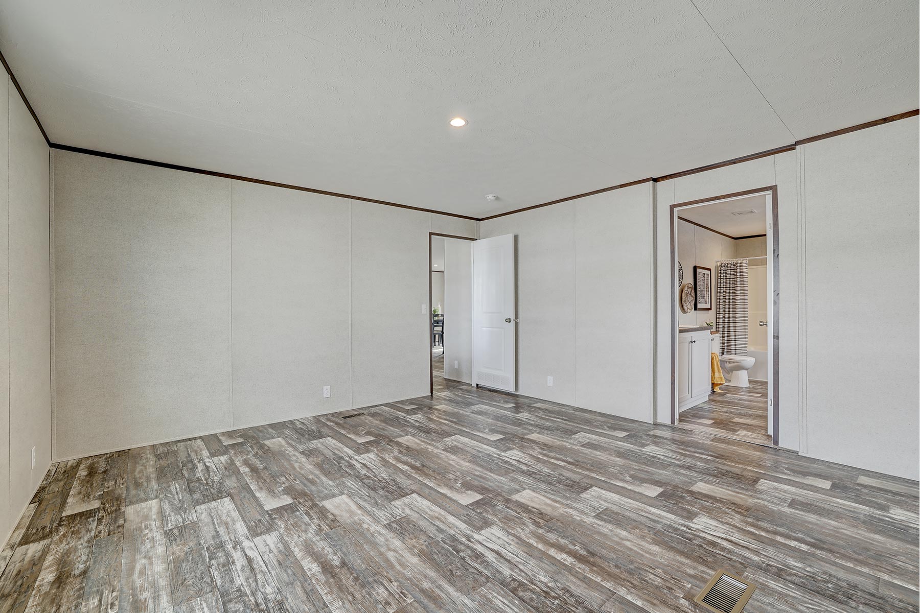 Empty room with light-colored walls and wood-style flooring. An open door reveals a bathroom, adding depth. The space feels bright and modern.