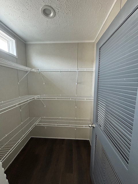 Compact walk-in pantry with white wire shelving on three walls, beige walls, dark wood flooring, and a light gray door. Natural light filters through a small window.