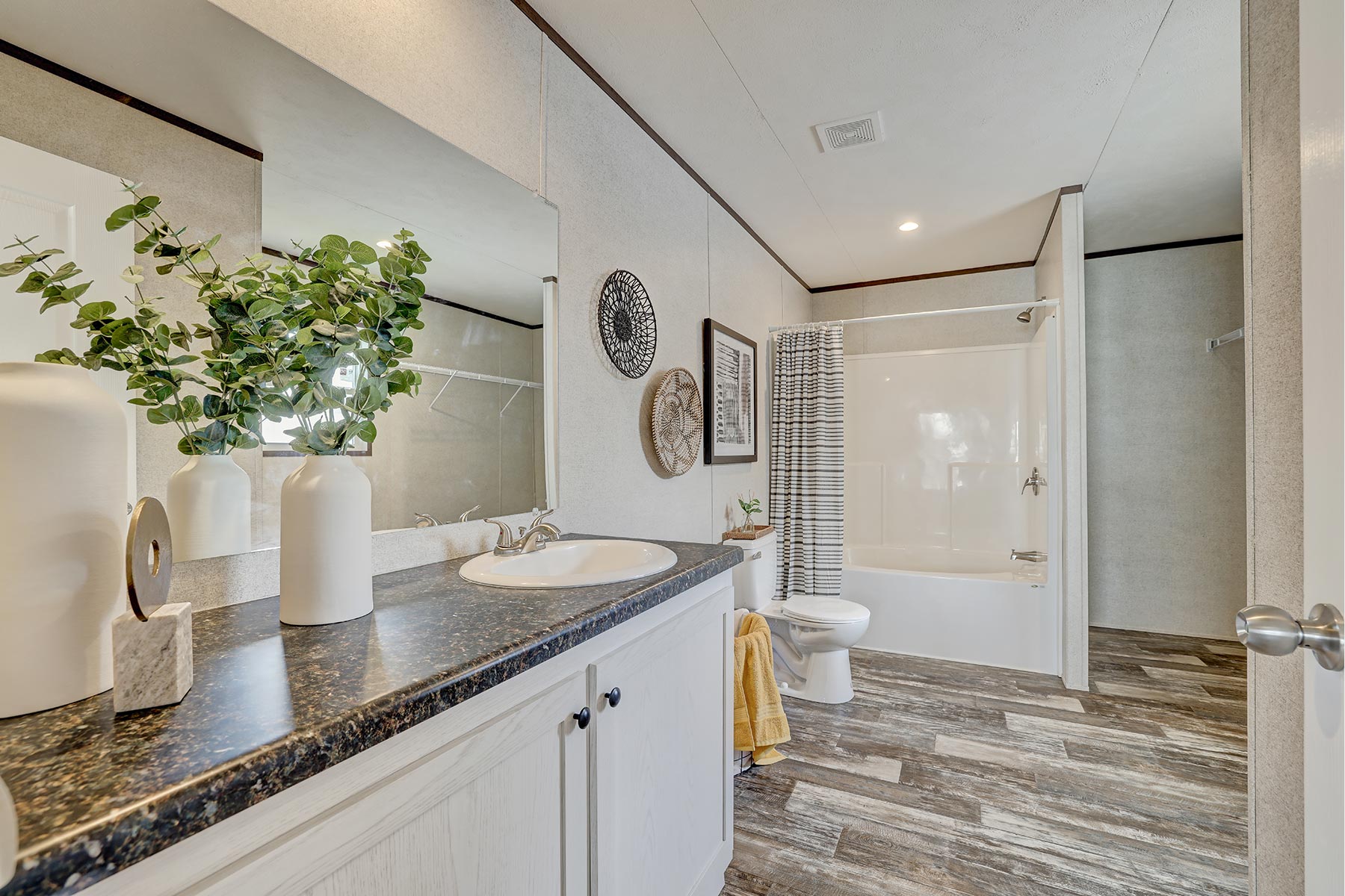 Modern bathroom with a dark countertop, white cabinets, and decorative plants. Wall art and yellow towel add warmth. Neutral tones create a calm vibe.