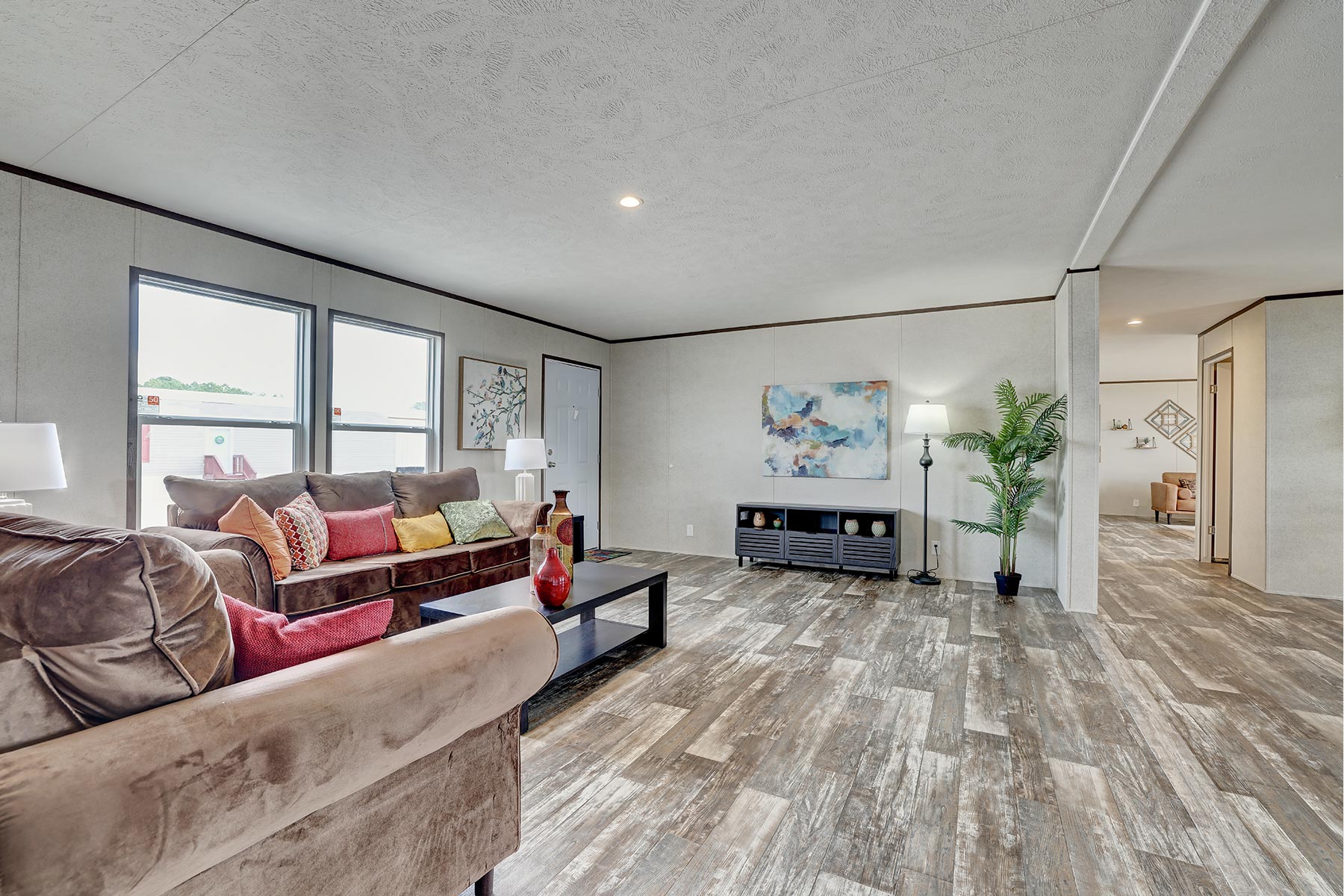 Spacious living room featuring a brown sofa with colorful cushions, wooden floor, abstract art on the walls, floor lamp, and potted plant for a cozy vibe.