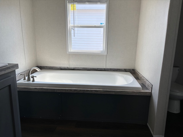 Spacious bathroom with a rectangular white bathtub set against light walls and a large window. Dark wood flooring and minimalist design evoke calmness.