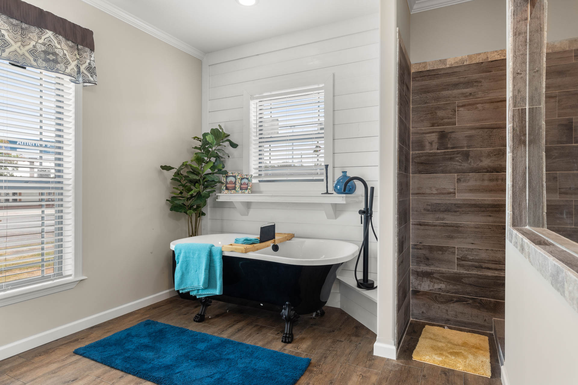 A modern bathroom features a black clawfoot tub with a wooden tray and turquoise towel, set against white shiplap walls. Nearby is a plant, a window with blinds, and a brown-tiled shower, creating a cozy, serene atmosphere.
