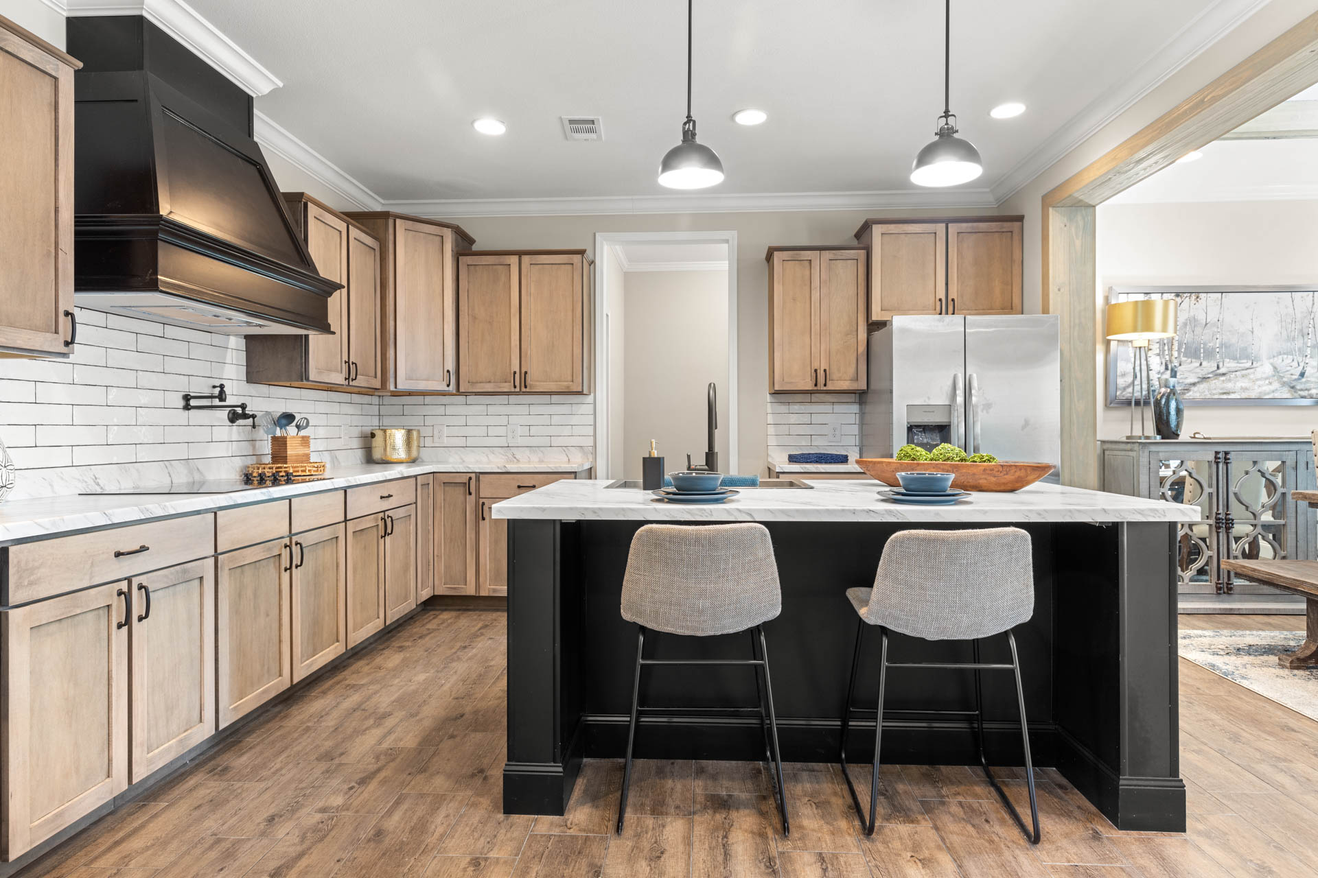 Modern kitchen with light wood cabinets, white subway tile backsplash, and stainless steel appliances. Black island with two gray chairs under pendant lights adds contrast. Warm and inviting atmosphere.