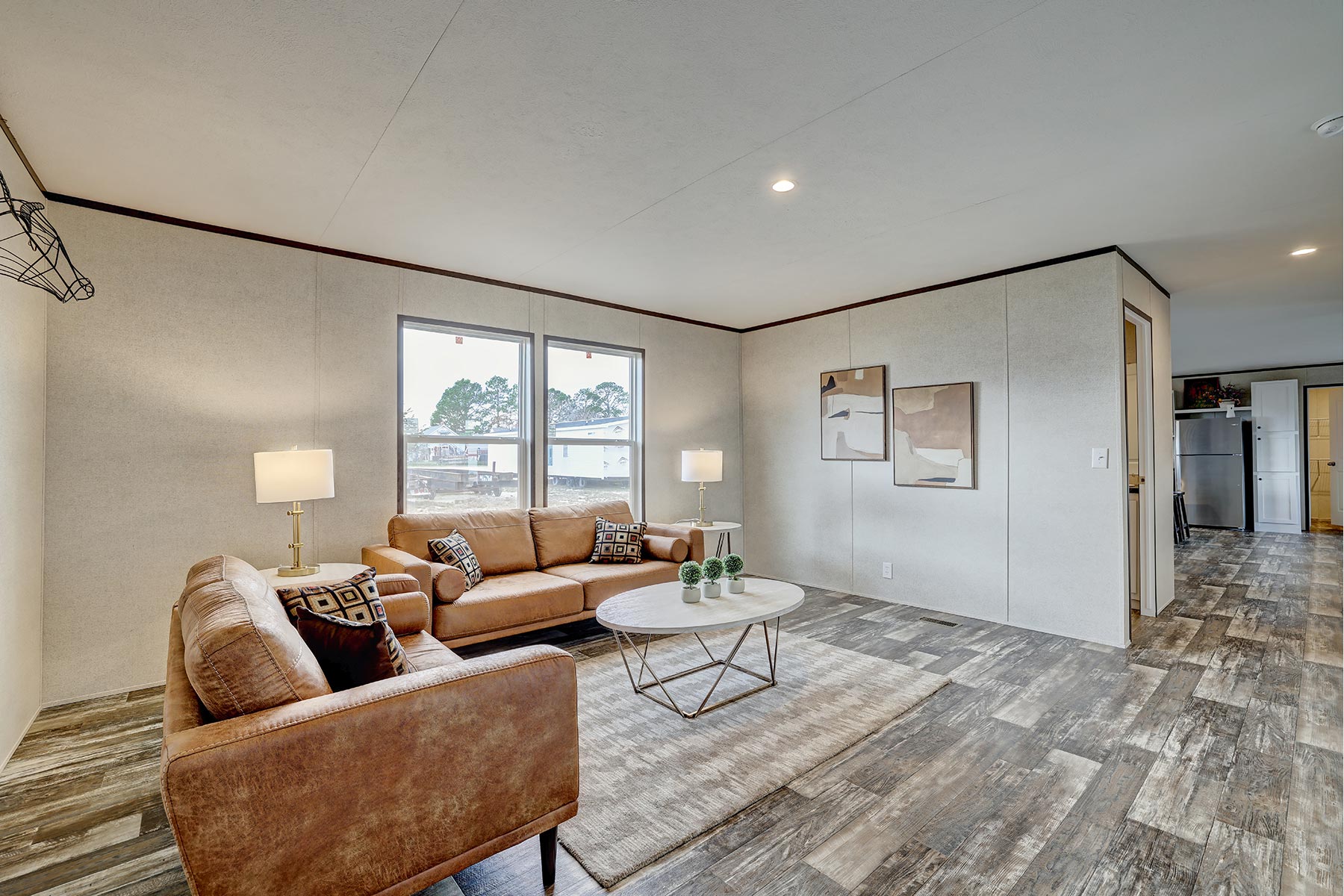 Spacious living room with tan sofas, patterned cushions, and a round coffee table. Bright lamps flank the sofa near windows. Art decorates the walls.